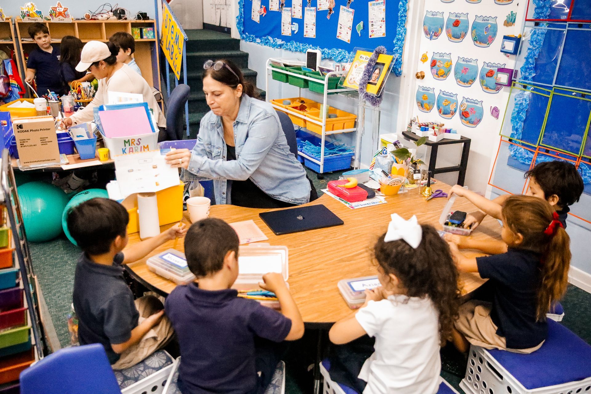 Children seated at a round table with a teacher, all engaged in an activity in a classroom.