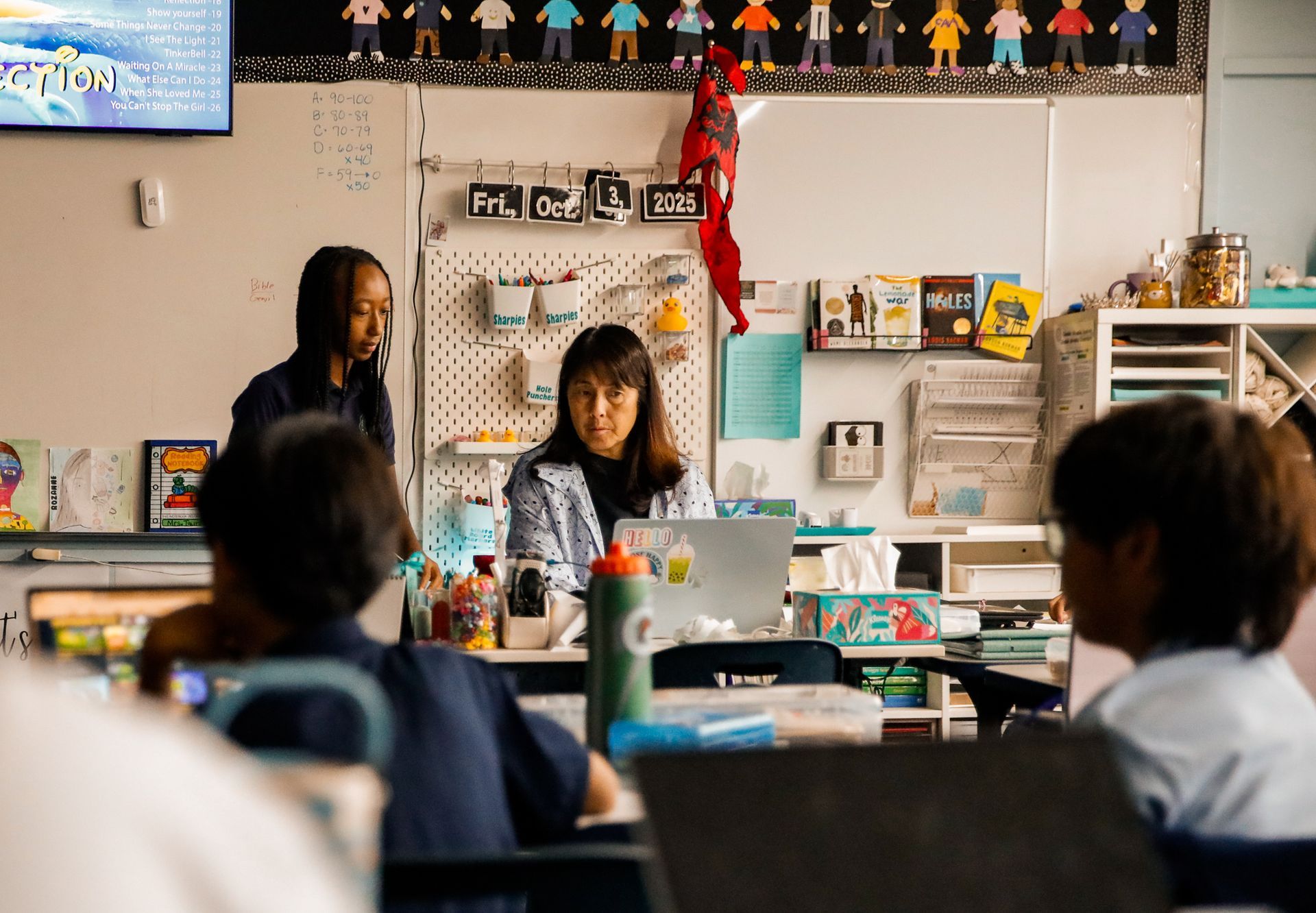 Two teachers in a classroom, one at a laptop. Children sit at desks, and the room has decorations.