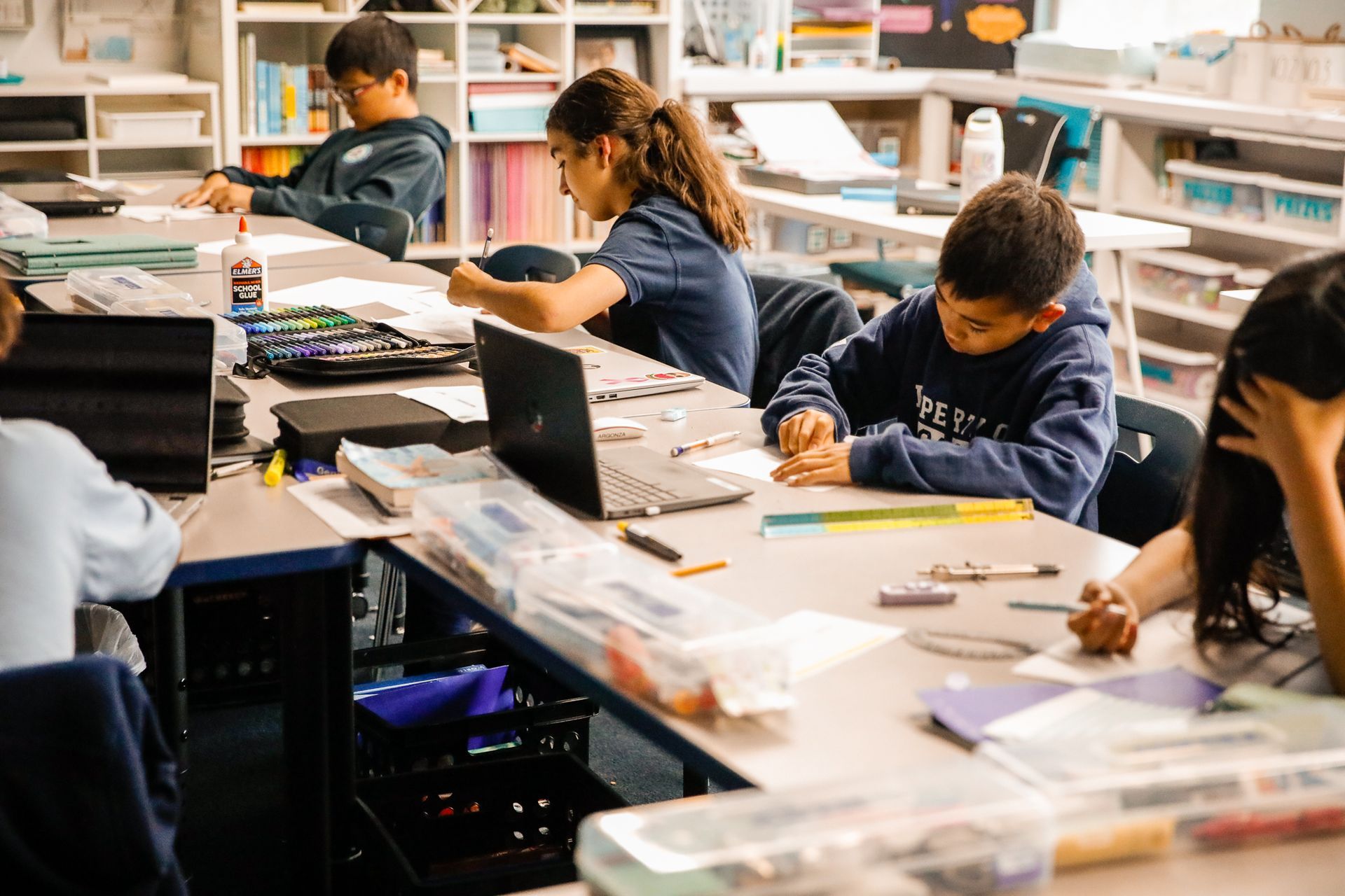 Students working at desks in a classroom, some using laptops and colorful supplies.