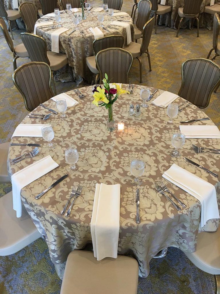 Round tables with beige patterned tablecloths and tan chairs set for a formal dining event.