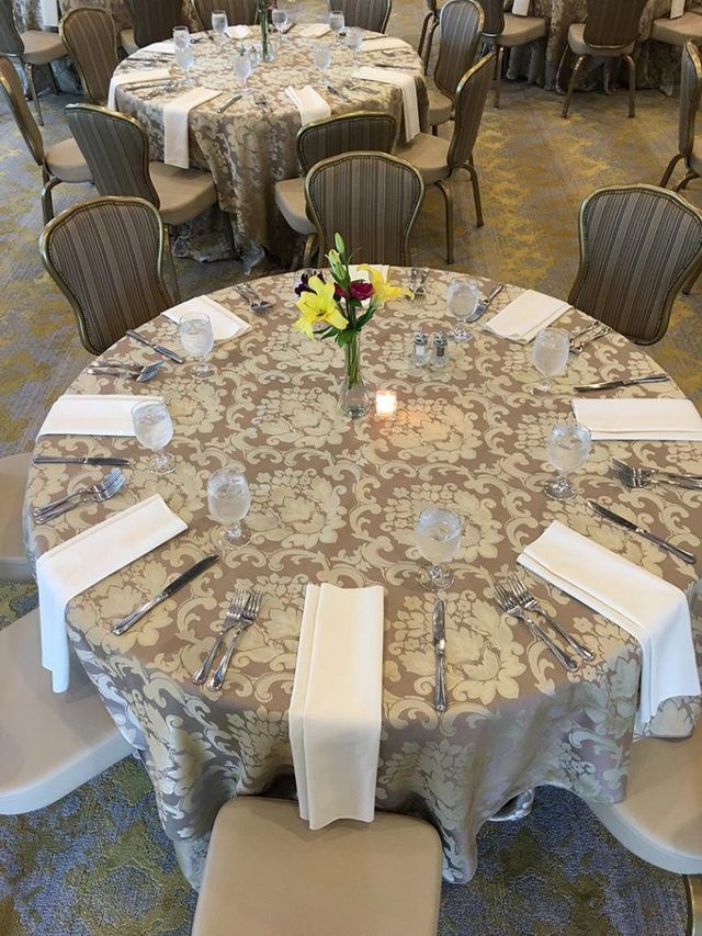 Round tables with beige patterned tablecloths and tan chairs set for a formal dining event.