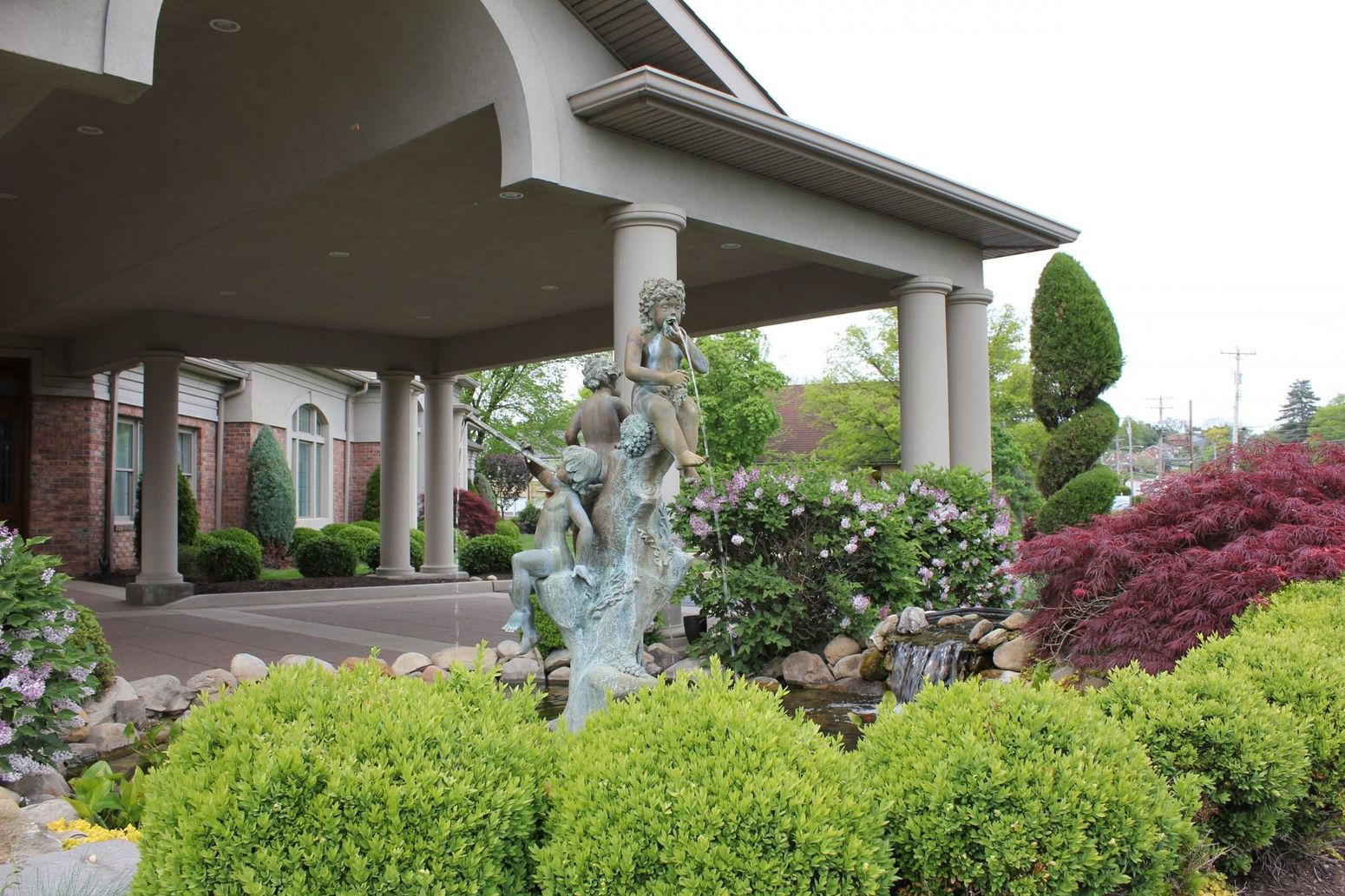 A stone fountain featuring cherub statues sits in front of a building with a pillared entrance, surrounded by shrubbery.