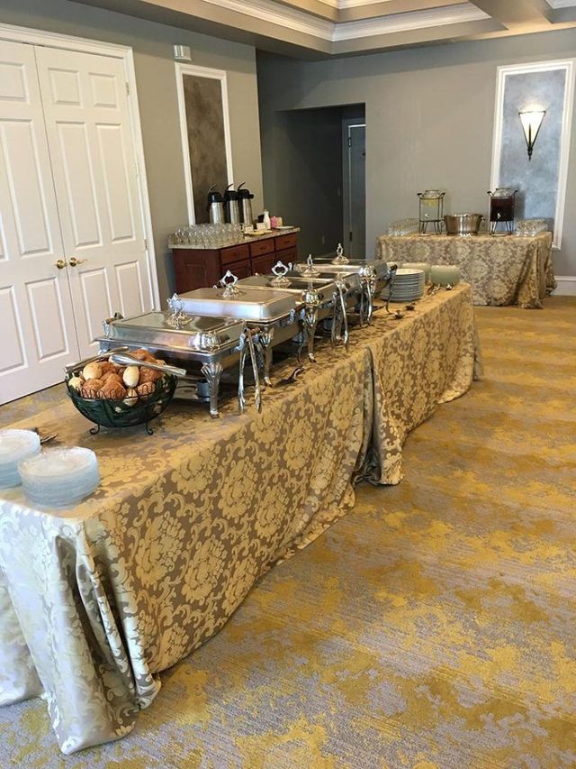 A buffet table set with chafing dishes, a bowl of rolls, and stacked plates in a room with patterned tablecloths.