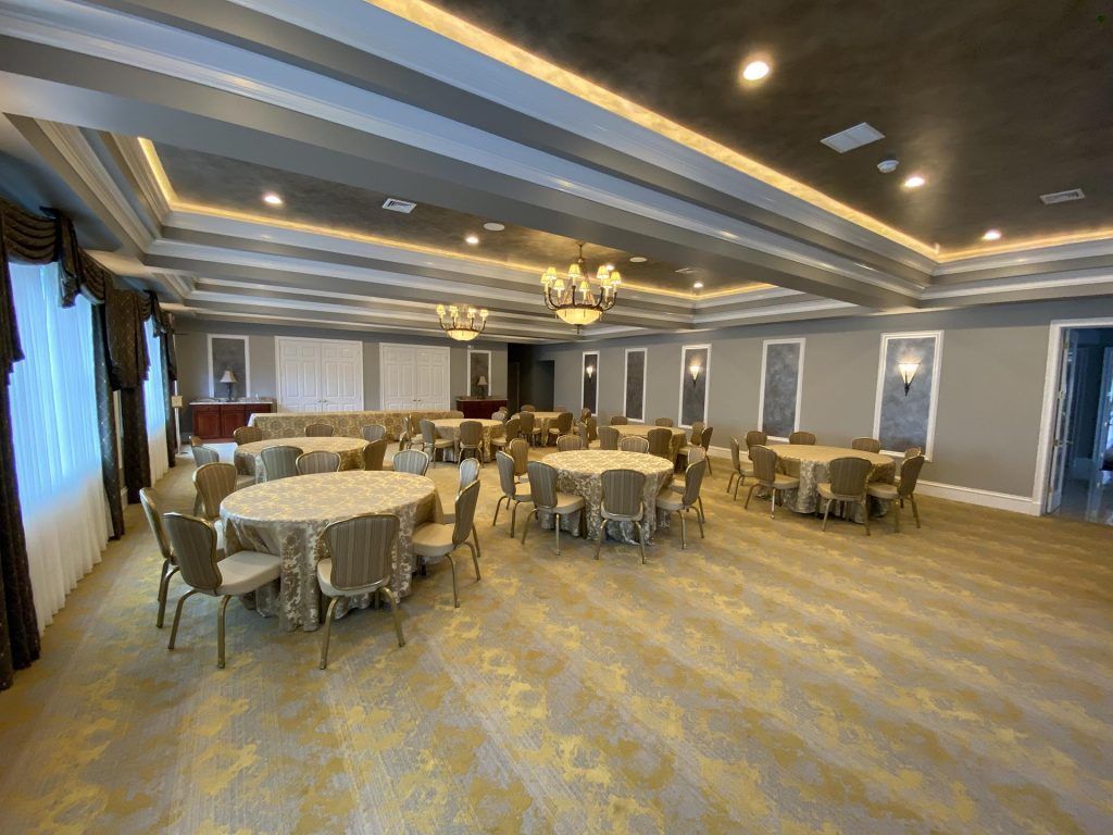 An empty banquet hall featuring round tables with tan linens, matching chairs, recessed tray ceilings, and gold carpet.