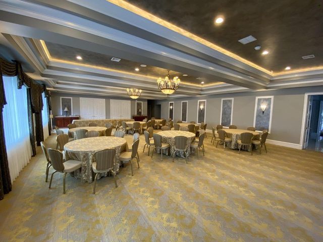 An empty banquet hall featuring round tables with tan linens, matching chairs, recessed tray ceilings, and gold carpet.