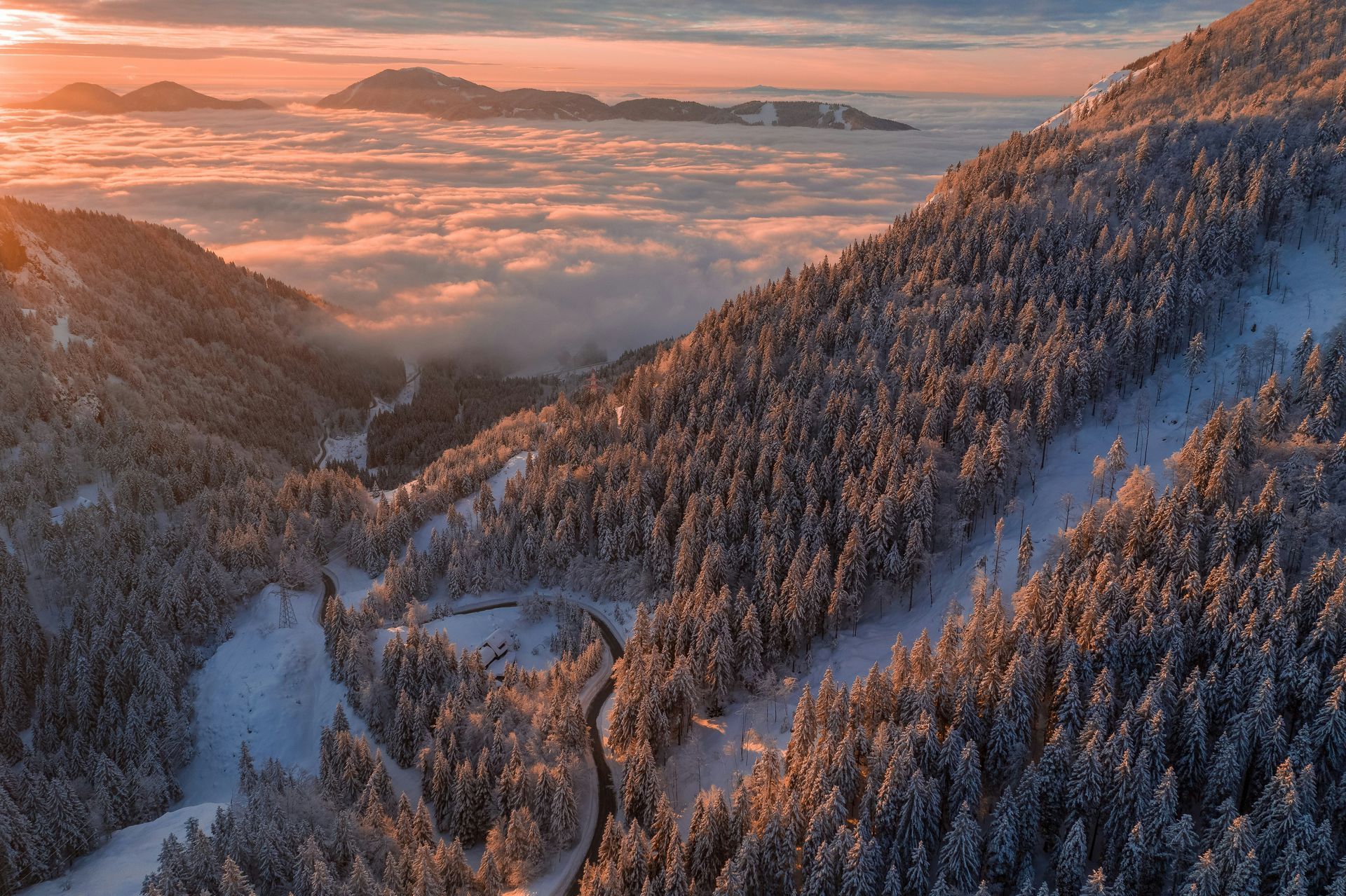 Snow-covered mountains at sunset. Valley with road winding through trees, above clouds. Warm light in the sky.