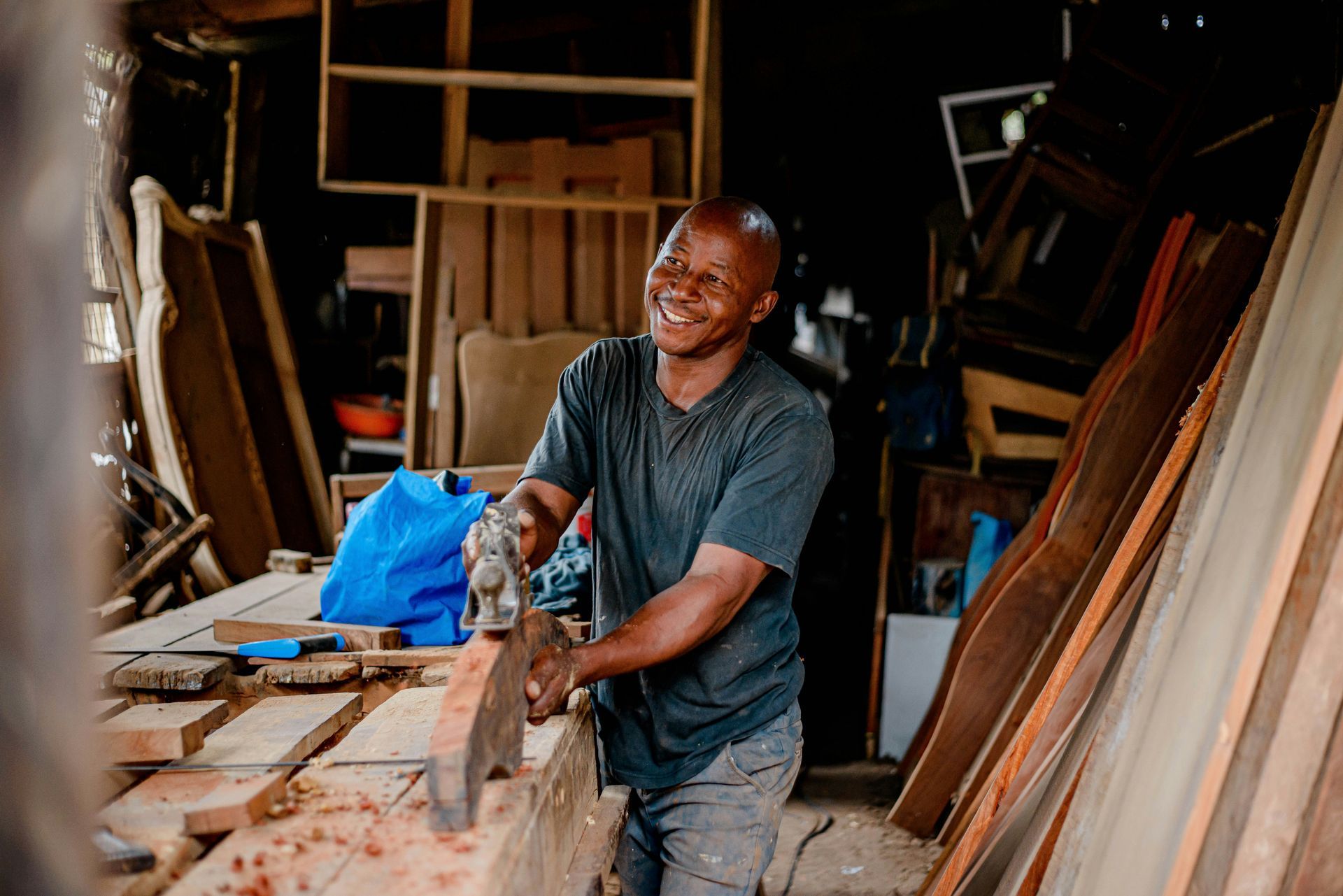 Man smiling while using a saw to cut wood in a workshop, surrounded by lumber and furniture.