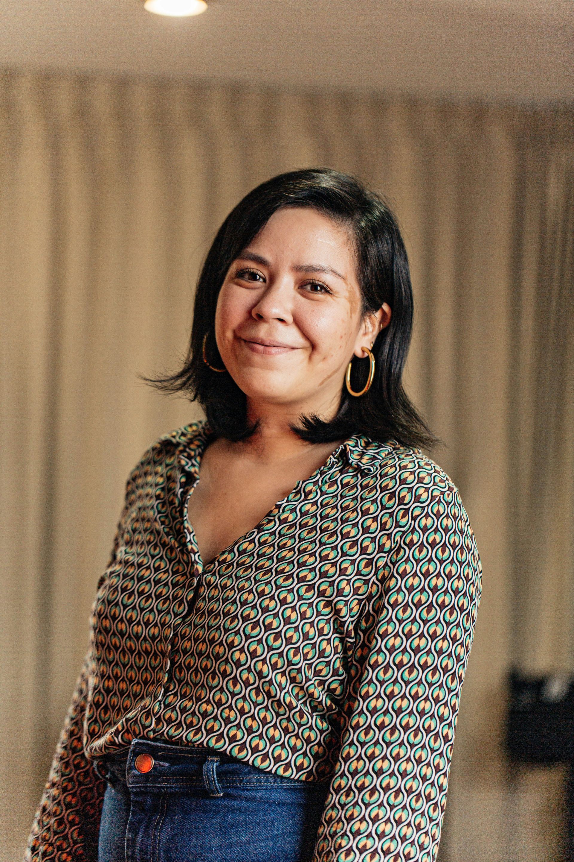 Woman smiling, wearing patterned shirt and hoop earrings, standing in front of curtains.