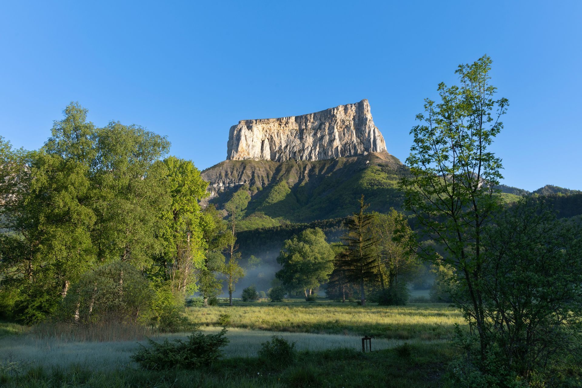 Flat-topped mountain rises above a green meadow. Trees frame the landscape under a clear blue sky.