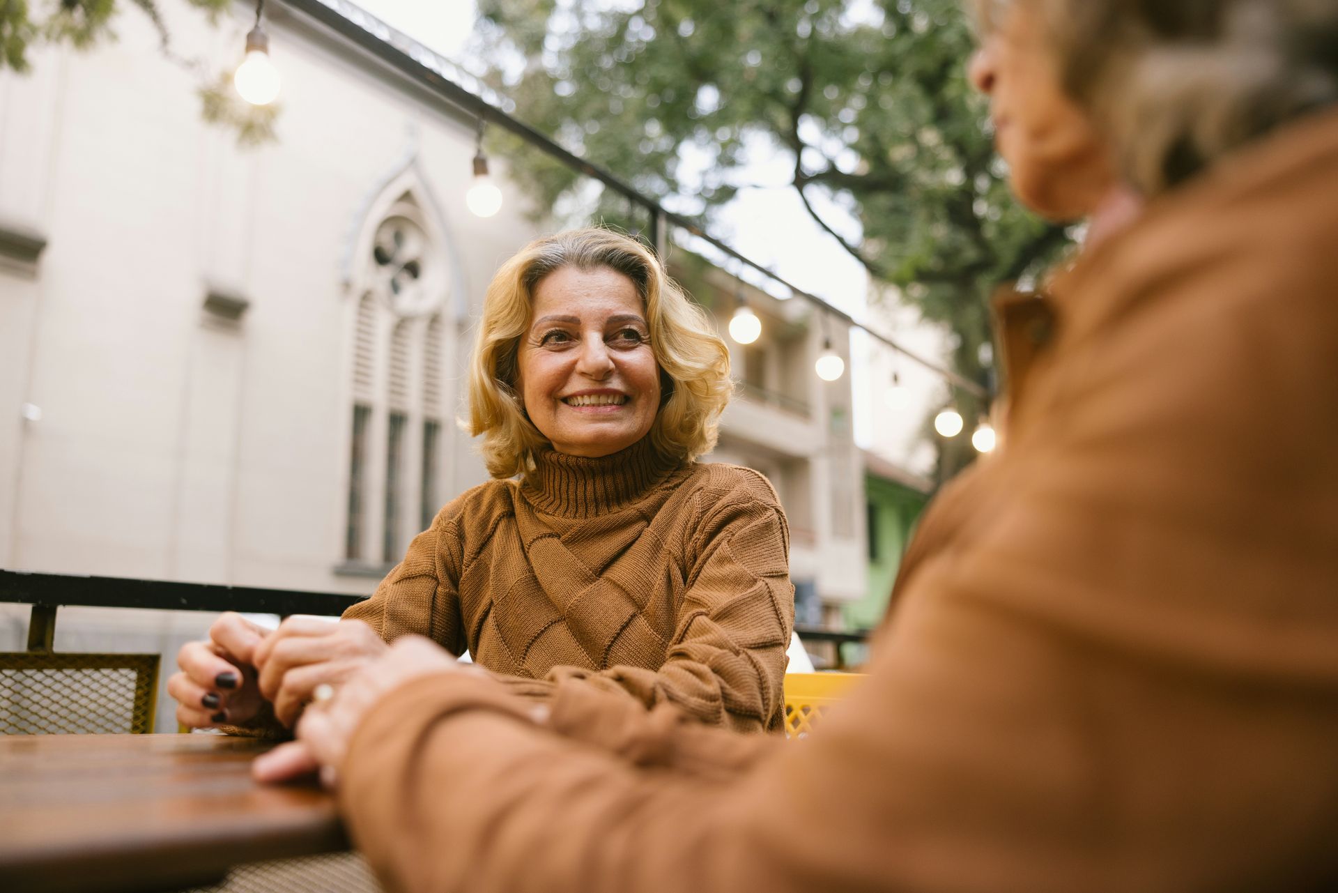 Woman smiles at another at outdoor cafe table; string lights and building in background.