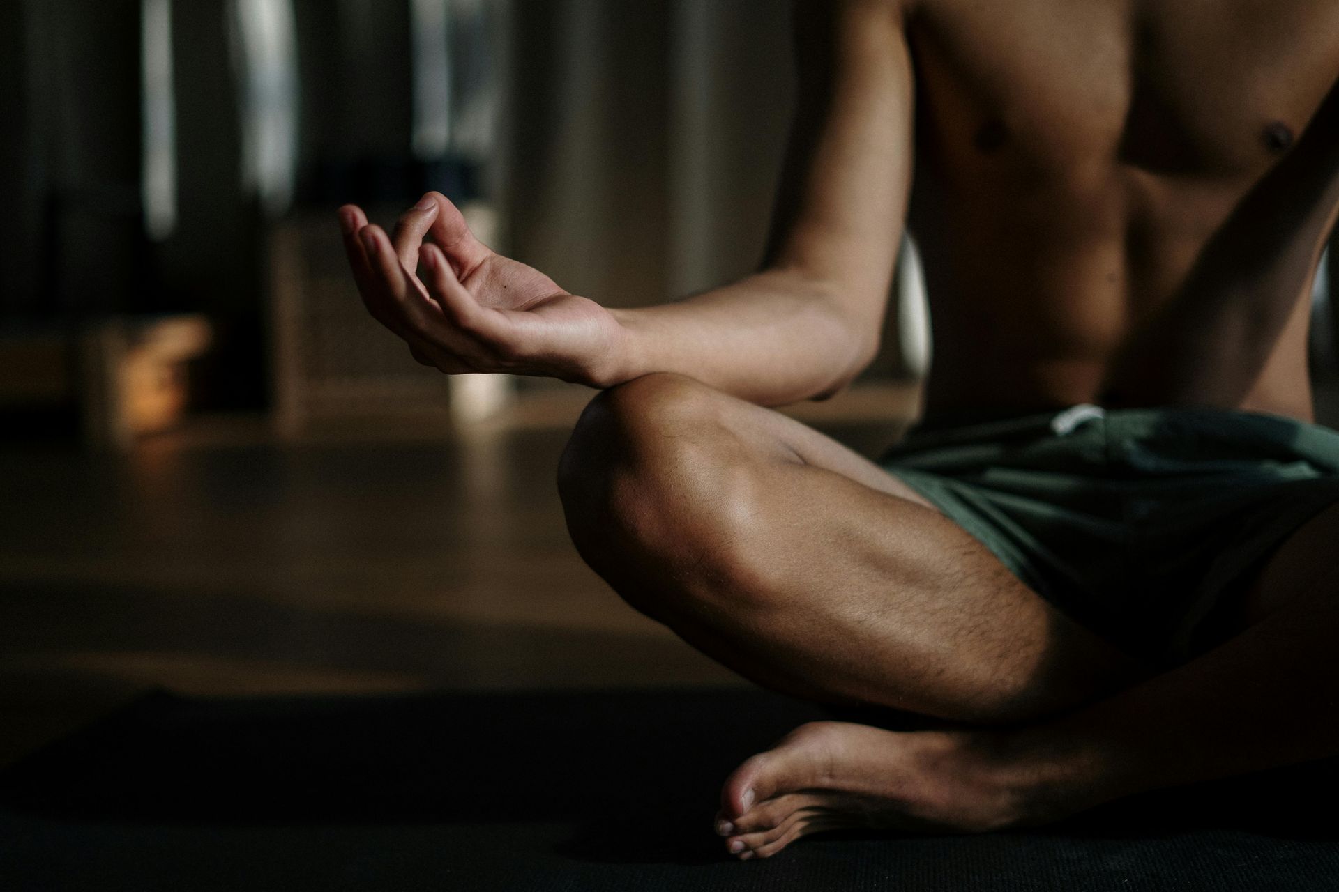 Man meditating cross-legged on a mat, hands in a mudra. Bare chest, shorts. Indoors.