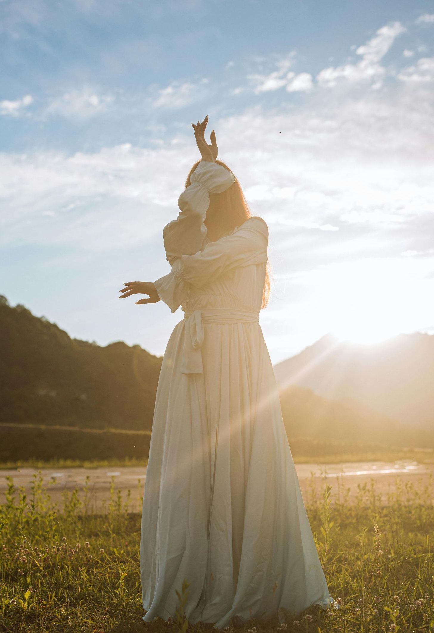 A woman in a white dress is standing in a field at sunset.