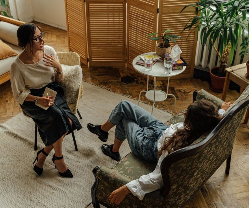 A woman is sitting in a chair talking to another woman who is laying on a couch.