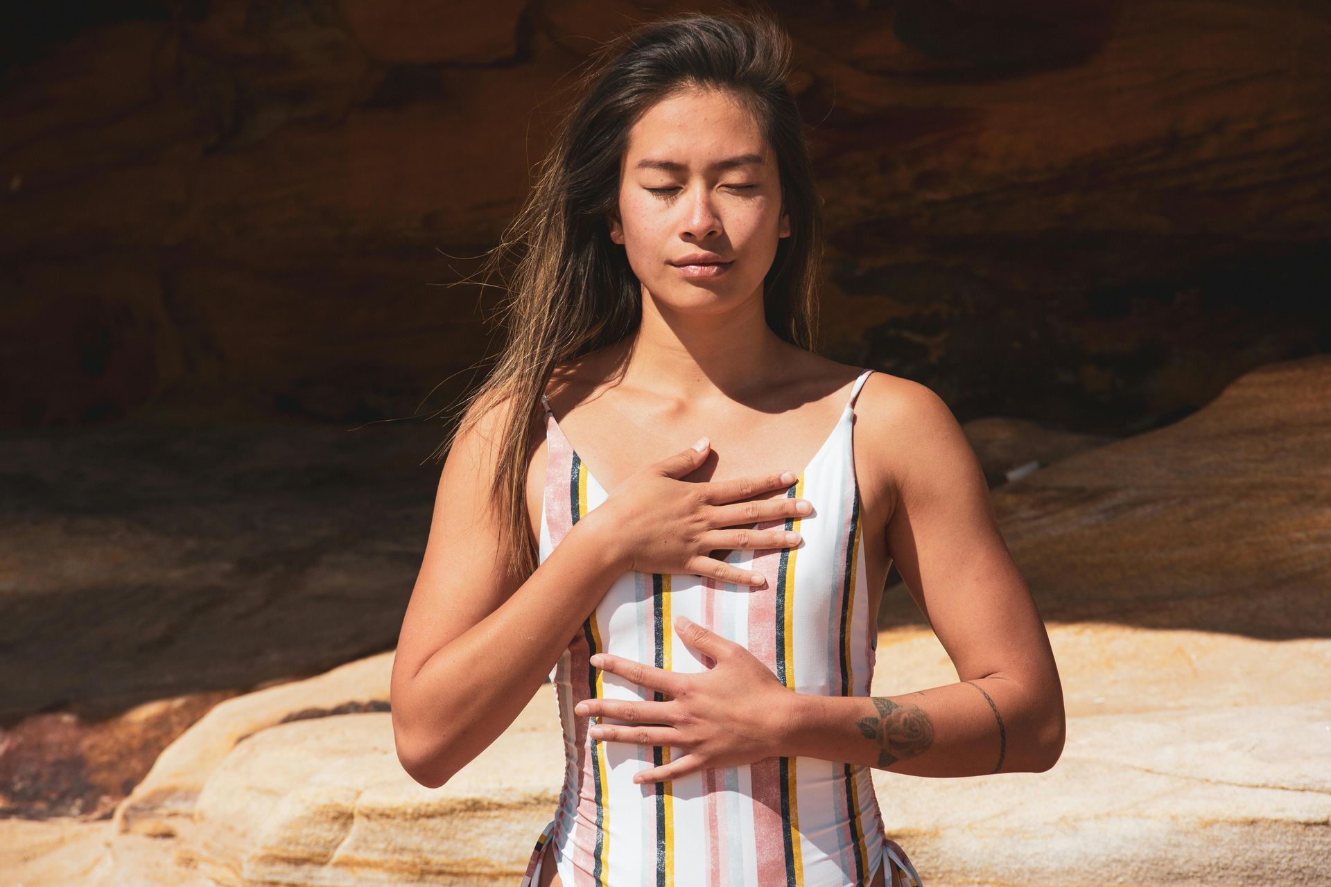 Woman in striped swimsuit, hands on chest, eyes closed, meditating near rock formations.