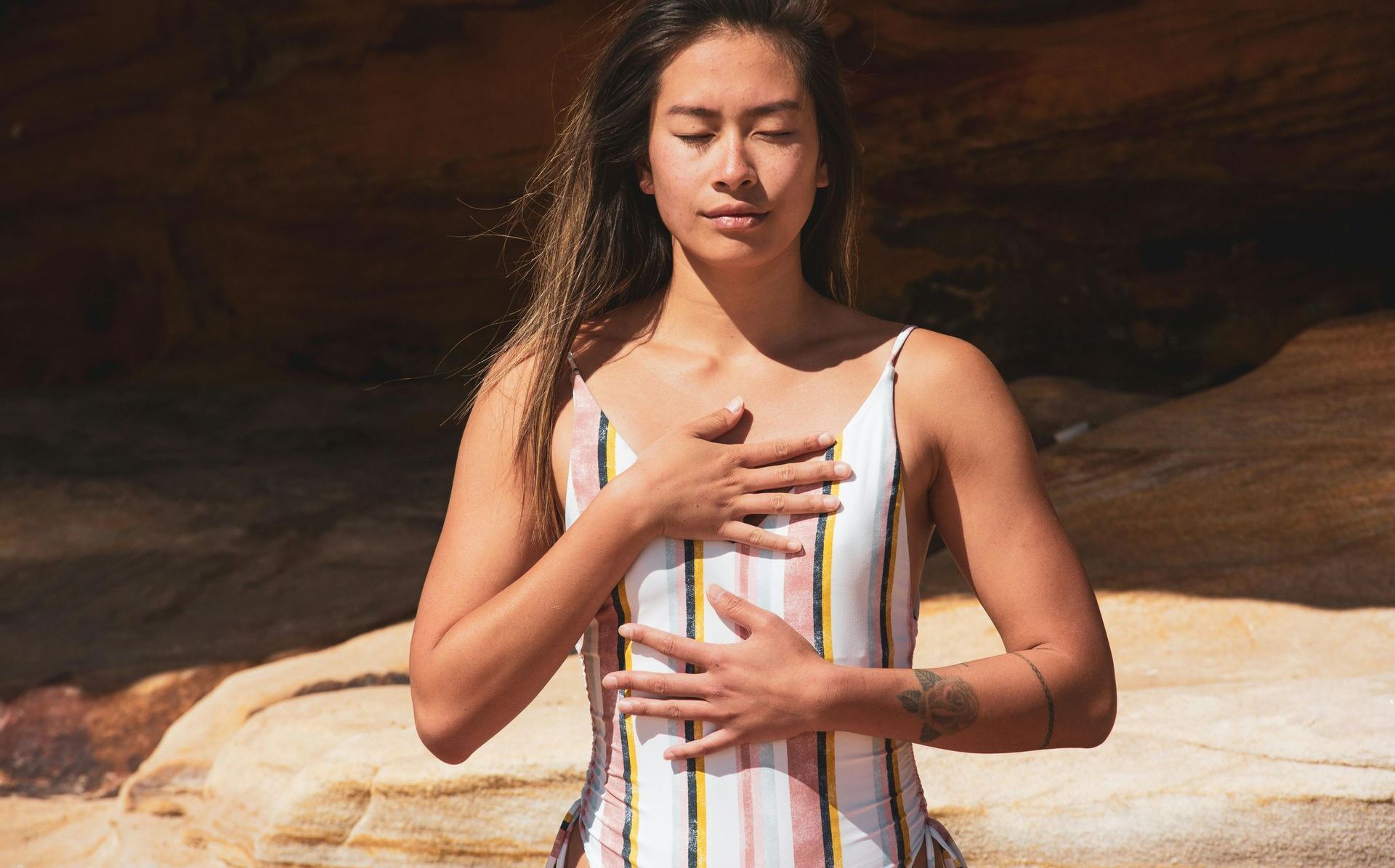 A woman with long brown hair sits in yoga seated pose in the desert, hands on heart and belly.