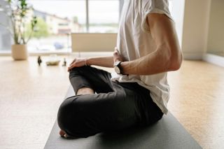 Person sitting cross-legged on a yoga mat in a light-filled room; wearing a white shirt and black pants; meditating.