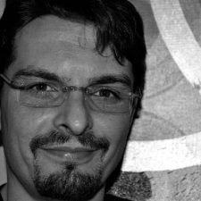 Uomo con occhiali e pizzetto sorridente, su uno sfondo sfocato e astratto.