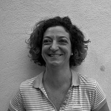 Una donna con i capelli ricci sorride, appoggiata a una parete ruvida, indossando una polo a righe.