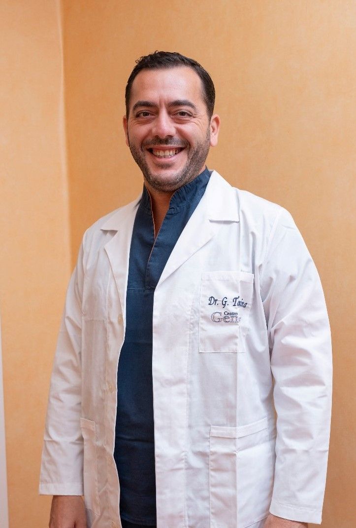 Un uomo sorridente con gli occhiali e un camice bianco, con le braccia incrociate, all'aperto.