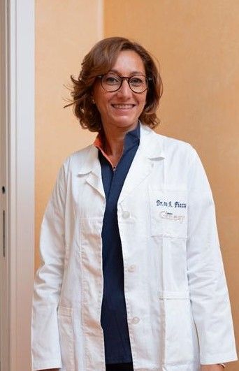 Foto in bianco e nero di una donna in camice, forse un medico, che sorride davanti a un fogliame. Indossa una sciarpa fantasia.