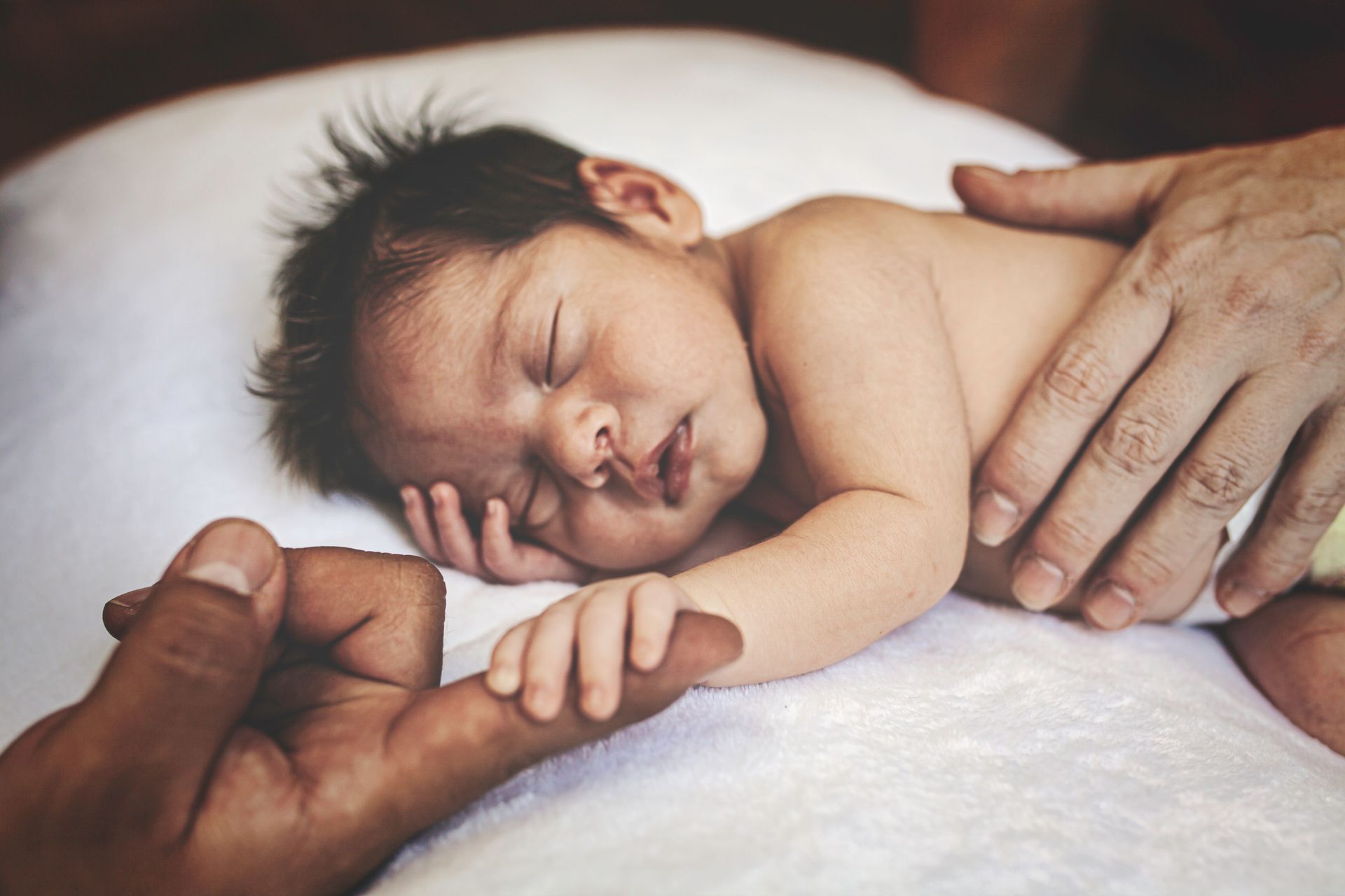 Un neonato dorme, con la mano appoggiata sulla fronte e il dito di un adulto. In un letto.