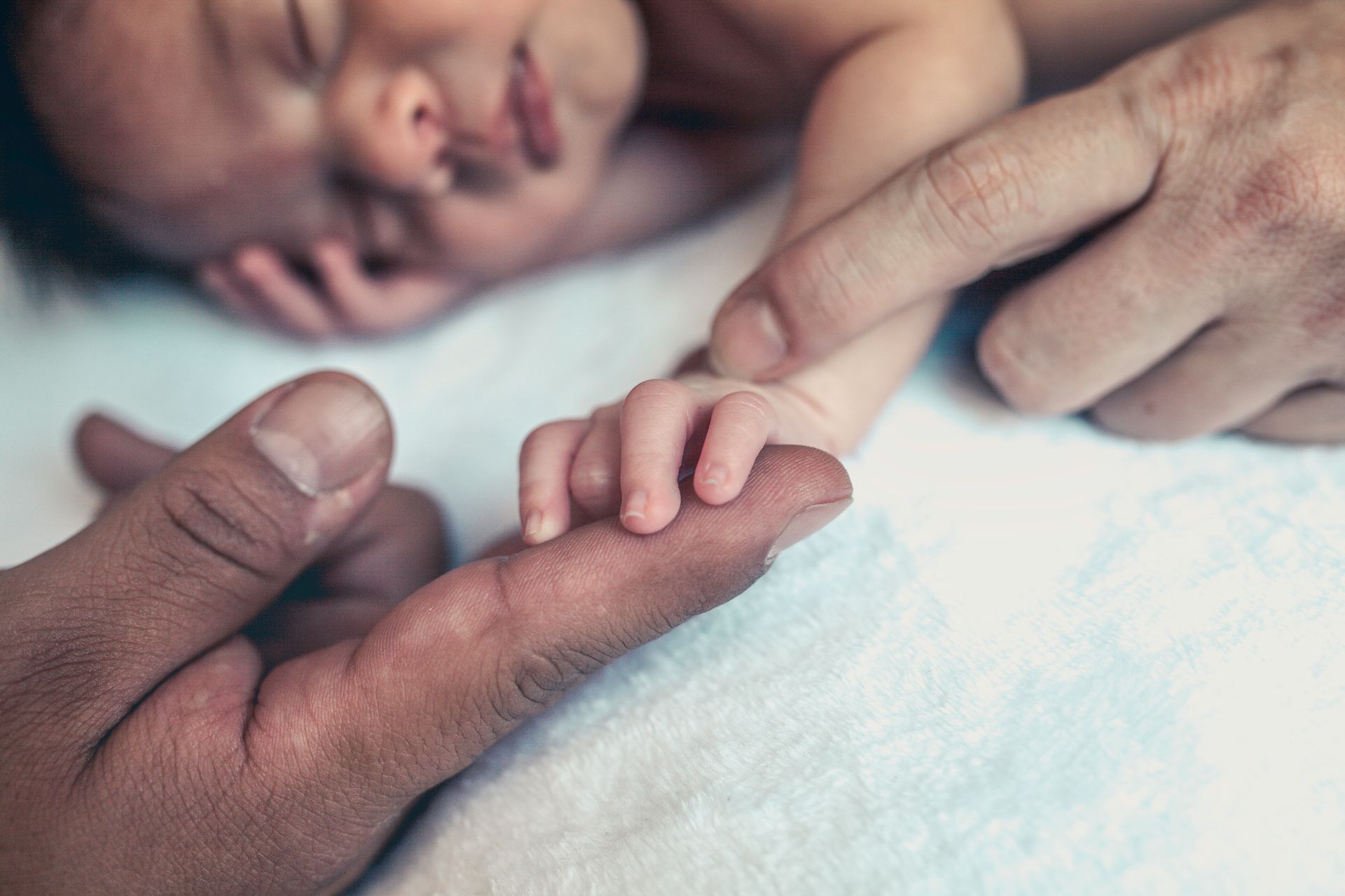 La piccola mano di un neonato addormentato afferra il dito di un adulto. Sullo sfondo una morbida coperta bianca.