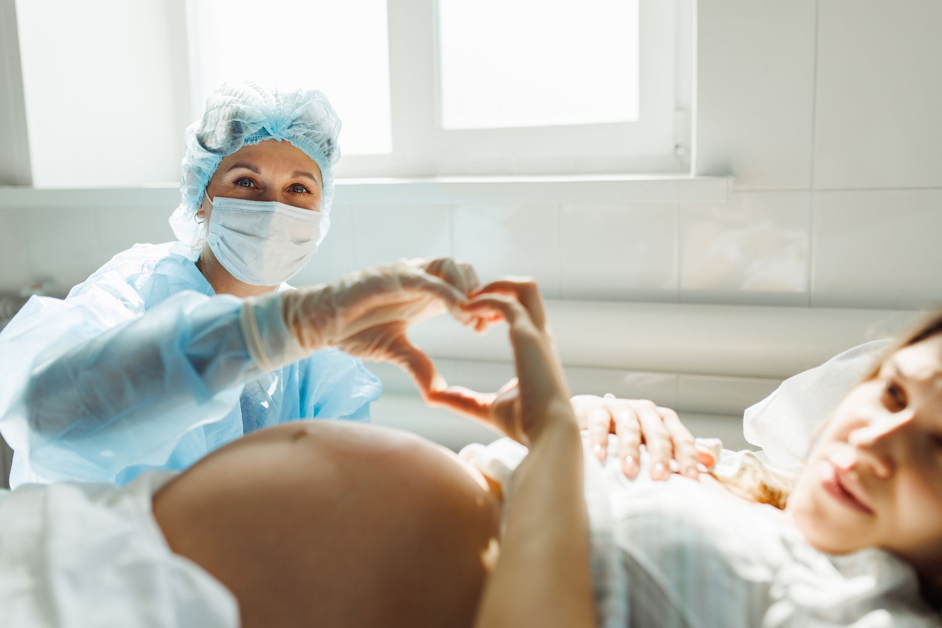 Una donna incinta in un letto d'ospedale sorride mentre un medico, che indossa una mascherina e una tuta chirurgica, disegna un cuore con le mani sulla pancia della donna.