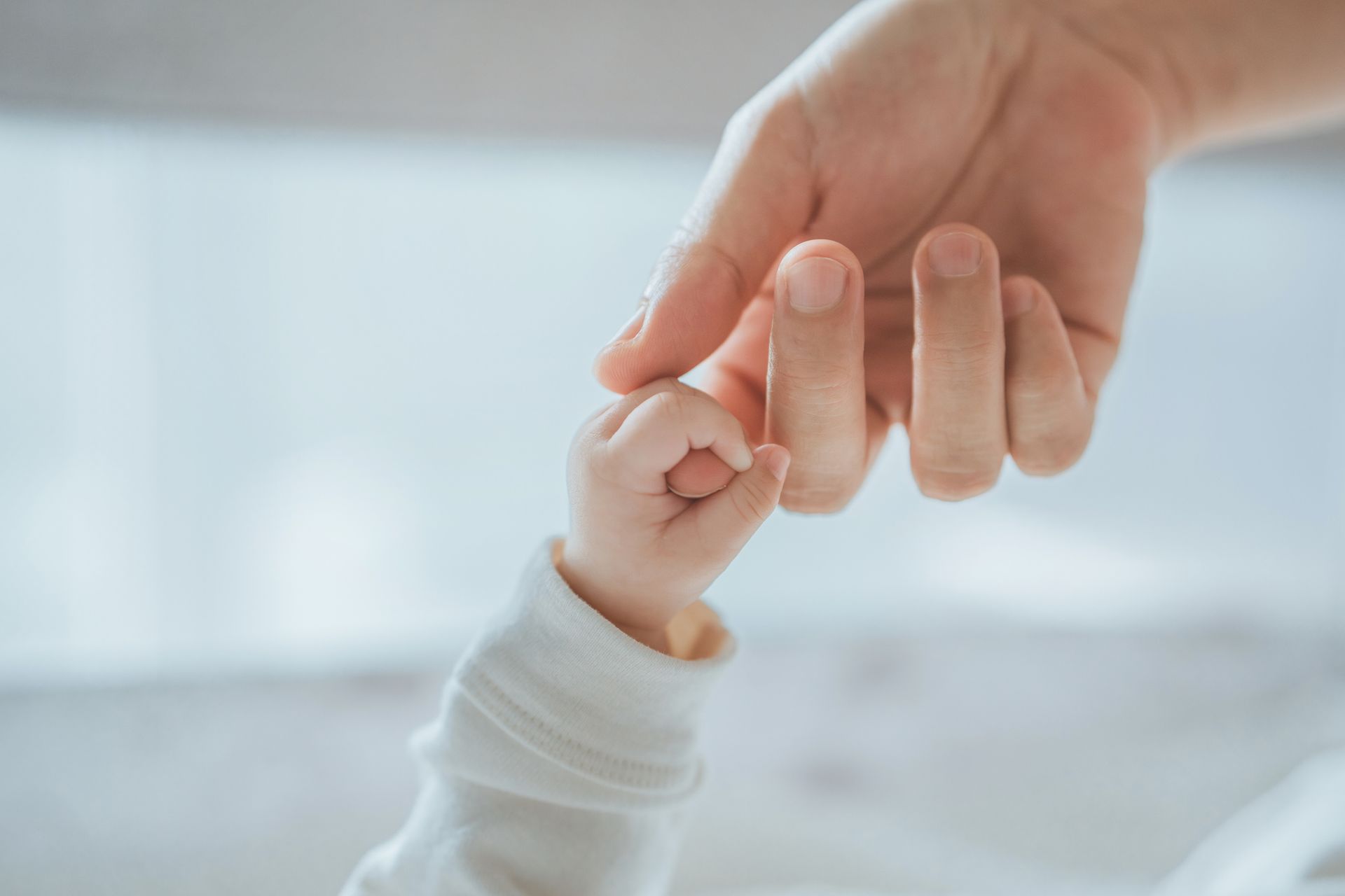 Una mano adulta tiene delicatamente la manina di un neonato, a simboleggiare la cura. Abiti e sfondo bianchi.