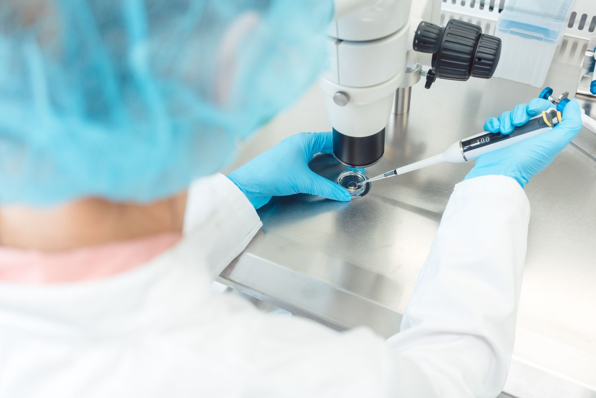 Un tecnico di laboratorio con guanti blu e berretto usa una pipetta sotto un microscopio. Si trovano in un ambiente di laboratorio luminoso e sterile.