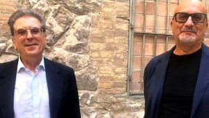 Due uomini in giacca e cravatta e con gli occhiali sono in piedi davanti a un muro di mattoni; uno sorride e l'altro guarda avanti.
