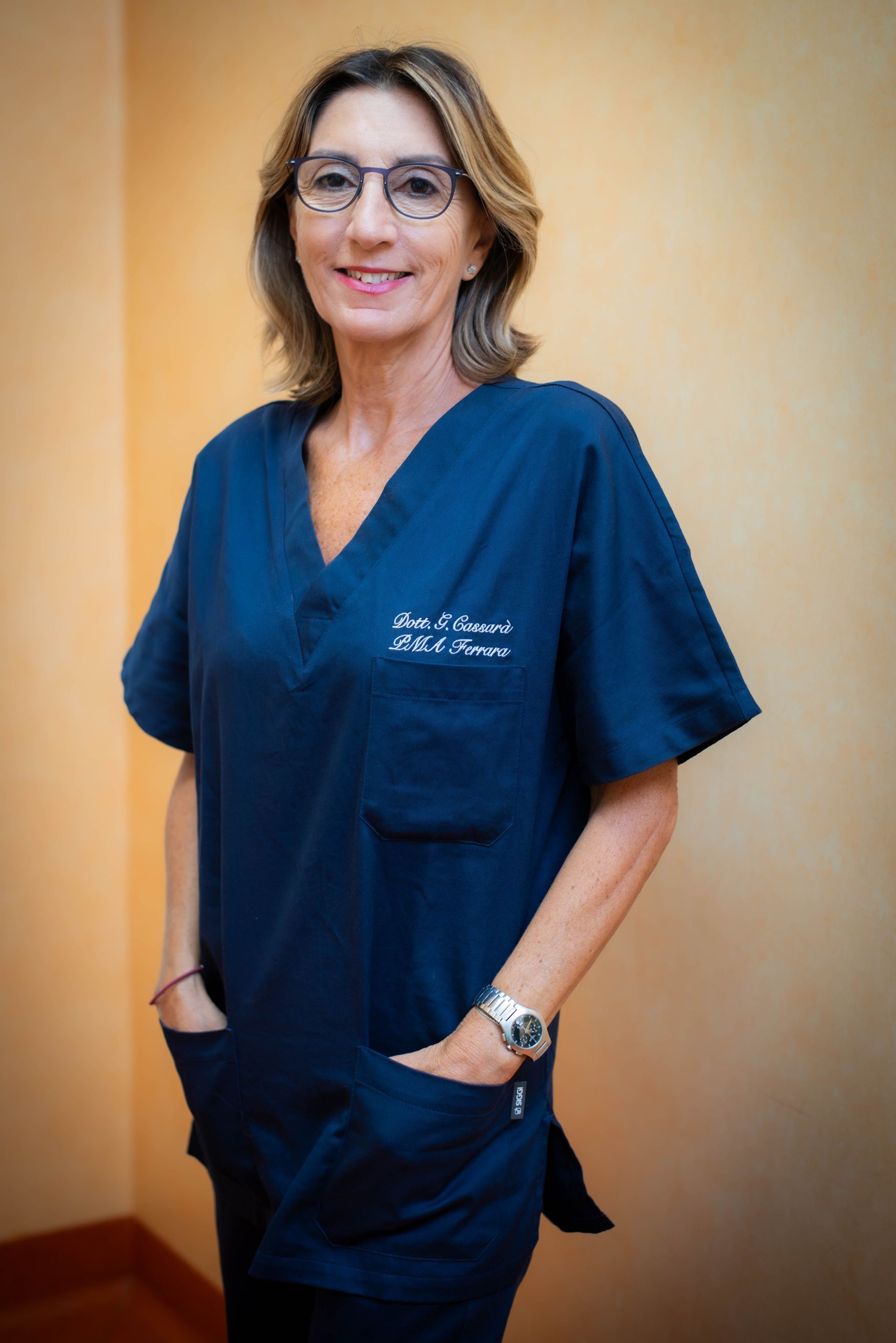 Donna sorridente in camice bianco, in piedi davanti a un fogliame verde.