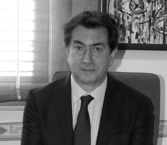 Un uomo in giacca e cravatta è seduto in un interno, rivolto verso la telecamera con un'espressione neutra. Sullo sfondo è appeso un dipinto.