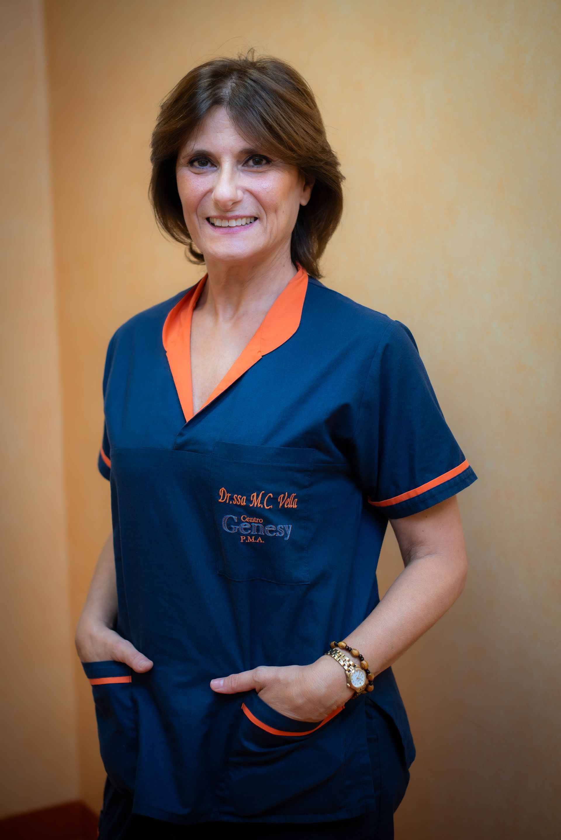 Donna con i capelli corti, sorridente, che indossa un cappotto bianco sopra un dolcevita nero e una collana, in piedi all'aperto.