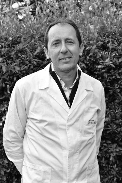 Un uomo in camice bianco sorride alla telecamera, in piedi davanti al fogliame verde.