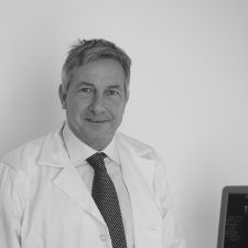 Un uomo che indossa un camice bianco, una cravatta e una camicia elegante sorride alla telecamera. Si trova in un ufficio, vicino al monitor di un computer.