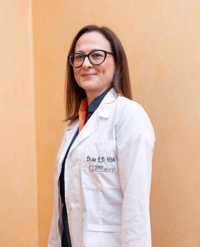 Una donna con gli occhiali, che indossa un camice da laboratorio, sorride davanti a uno sfondo verdeggiante.