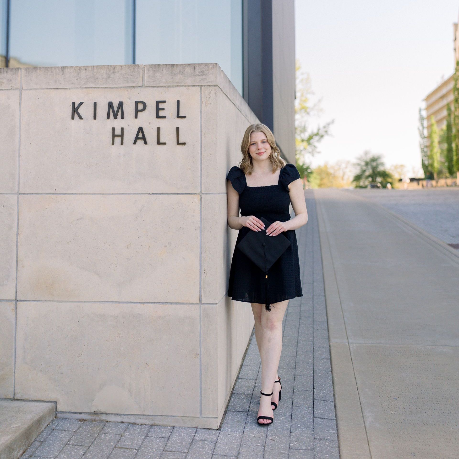 A woman in a black dress is leaning against a wall that says kimpel hall
