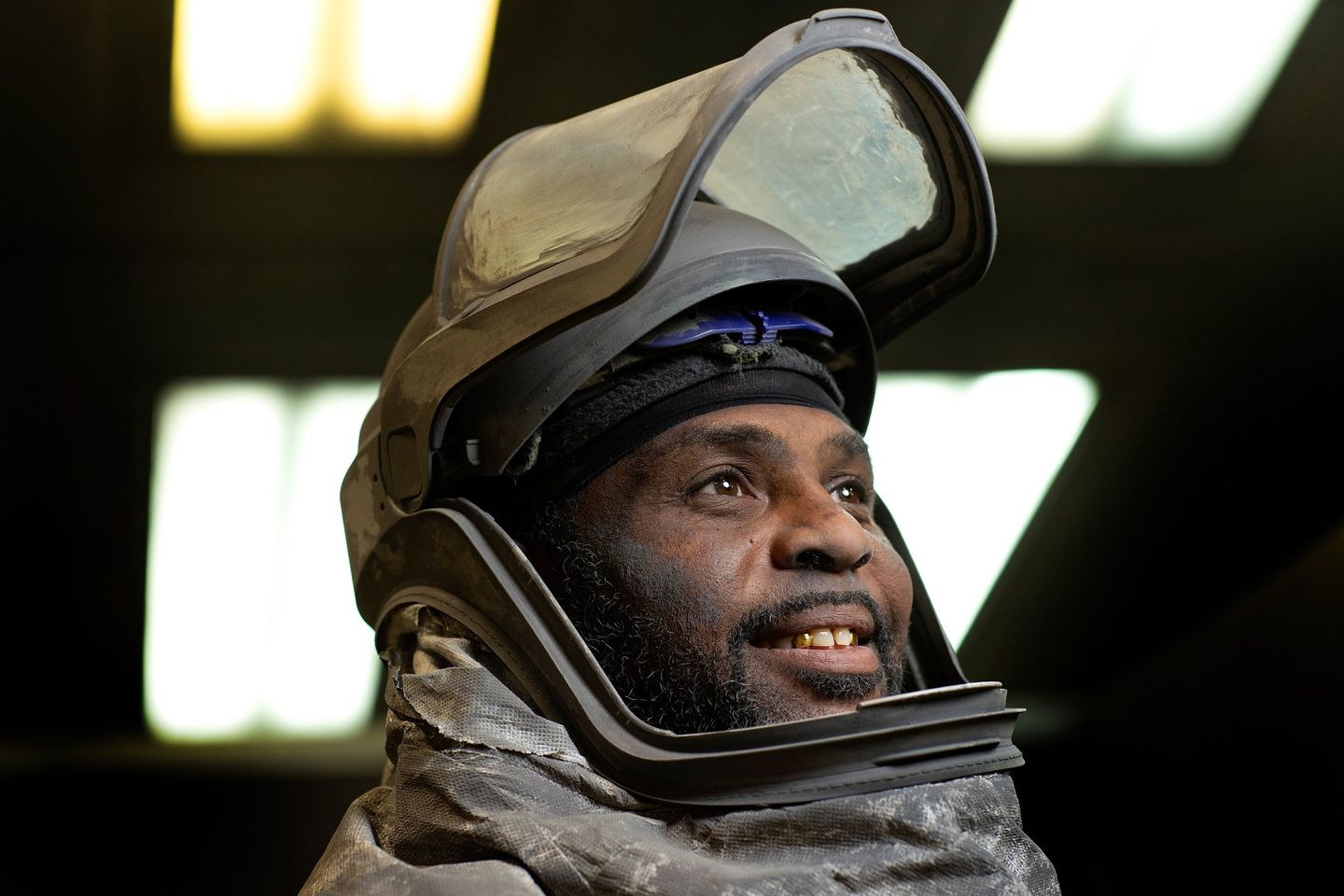 A man wearing a full-body suit used in industrial applications is posing for a photo. He is not looking at the camera. His face shield is up so his face is visible. He looks happy. 