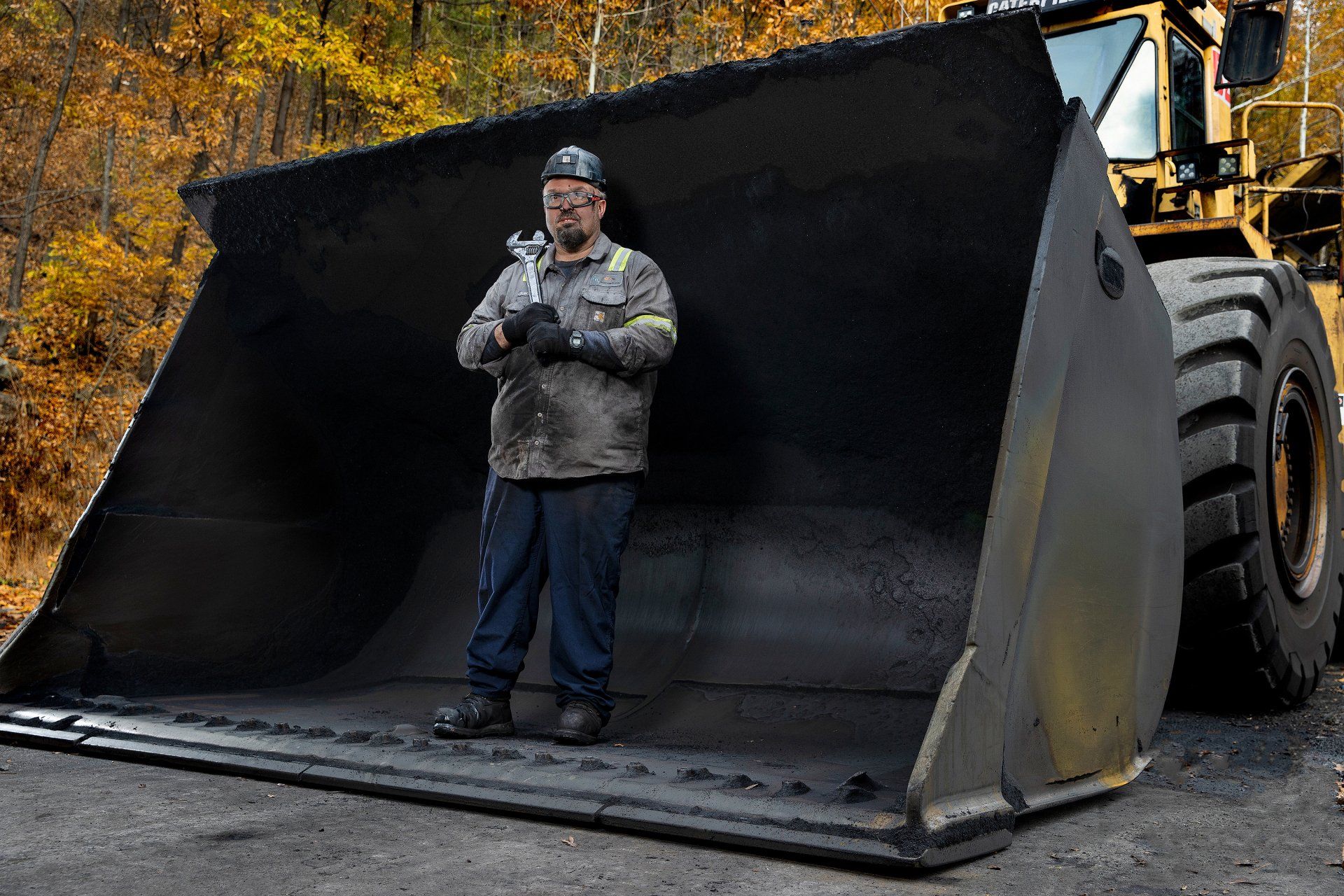 A construction worker stands inside the scoop of a large mover, which is larger than the man. 