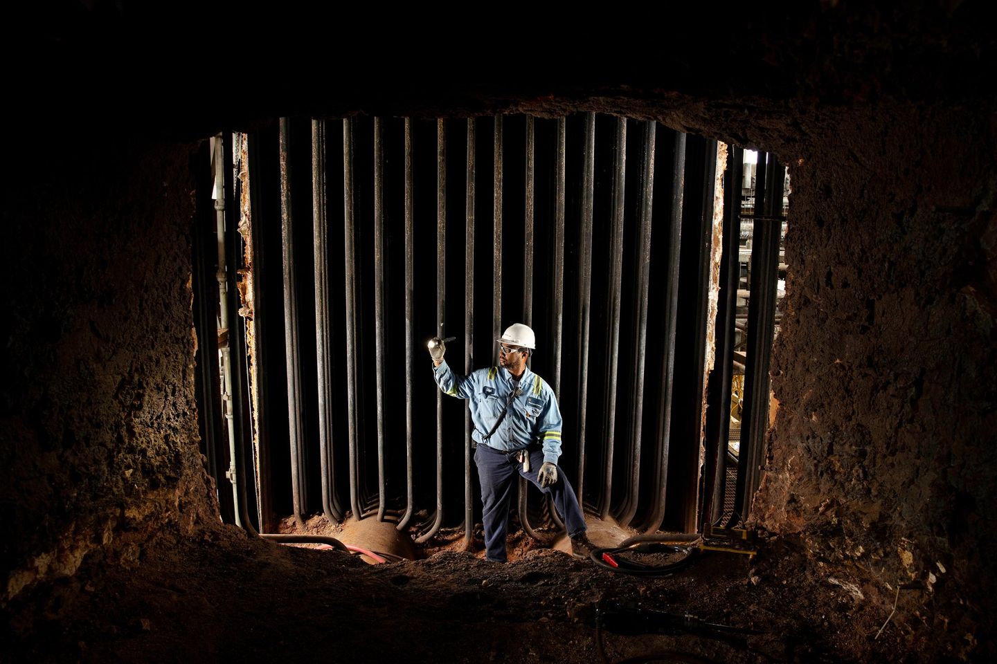 A male employee has his photo taken at the entrance to a dirty industrial furnace. The man is holding a flashlight and appears to be inspecting the furnace. 
