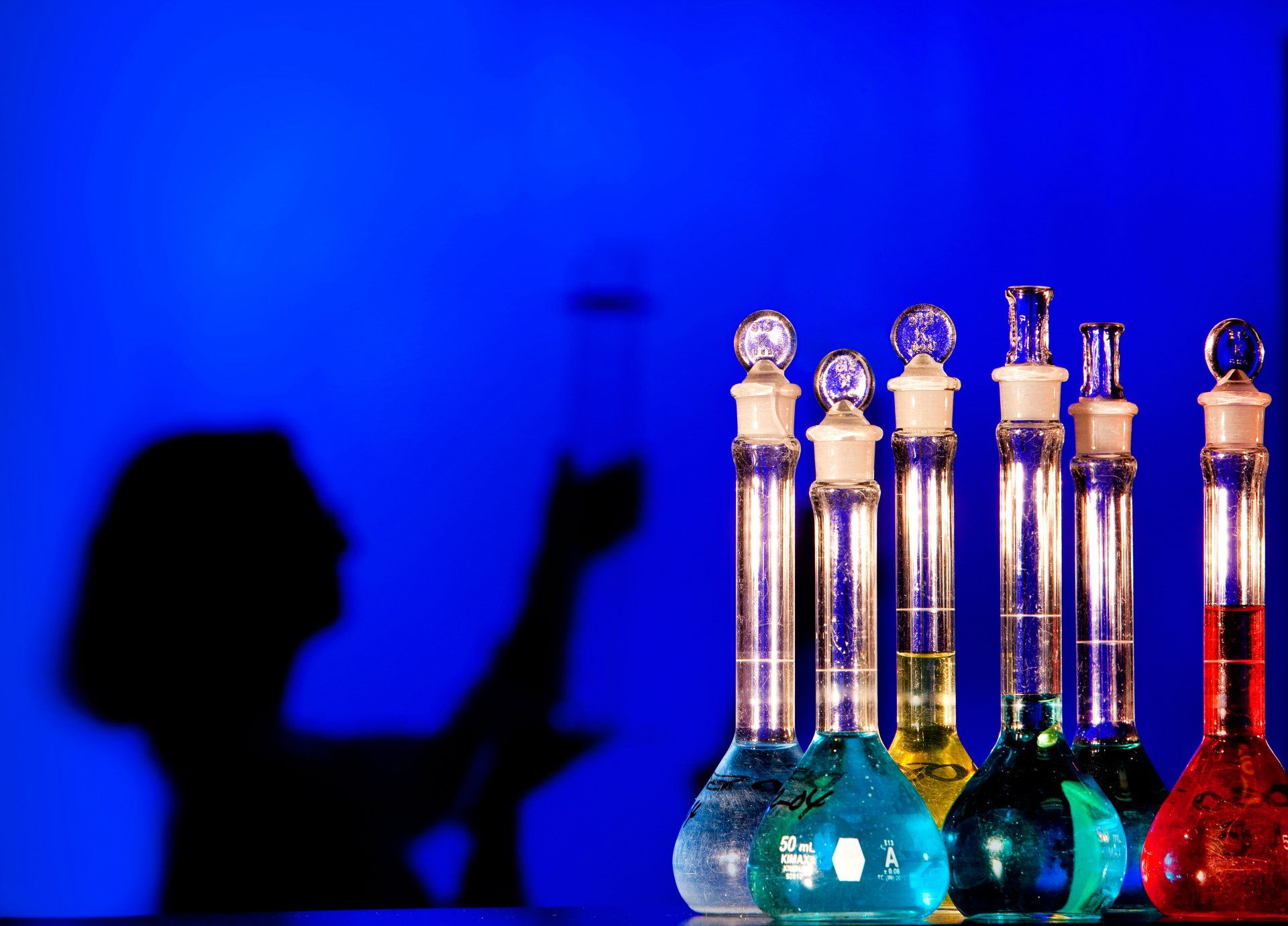 A woman laboratory scientist is silhouetted behind vibrant vials of liquids in her research lab.
