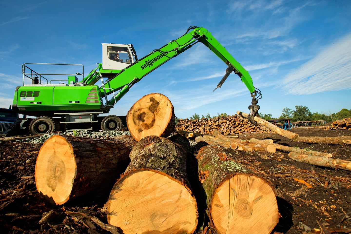 An male operator in a lumber mill uses a machine with a large jaw to move logs around the yard. 