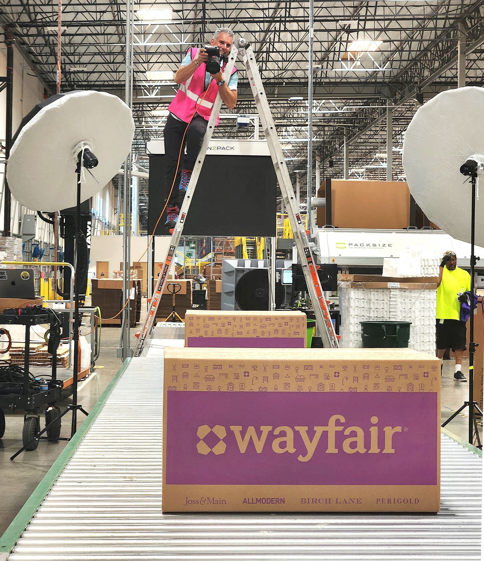 Commercial manufacturing photographer takes photos while he stands at the top of a ladder. The photographer is taking pictures of boxes in a logistics and warehouse facility.