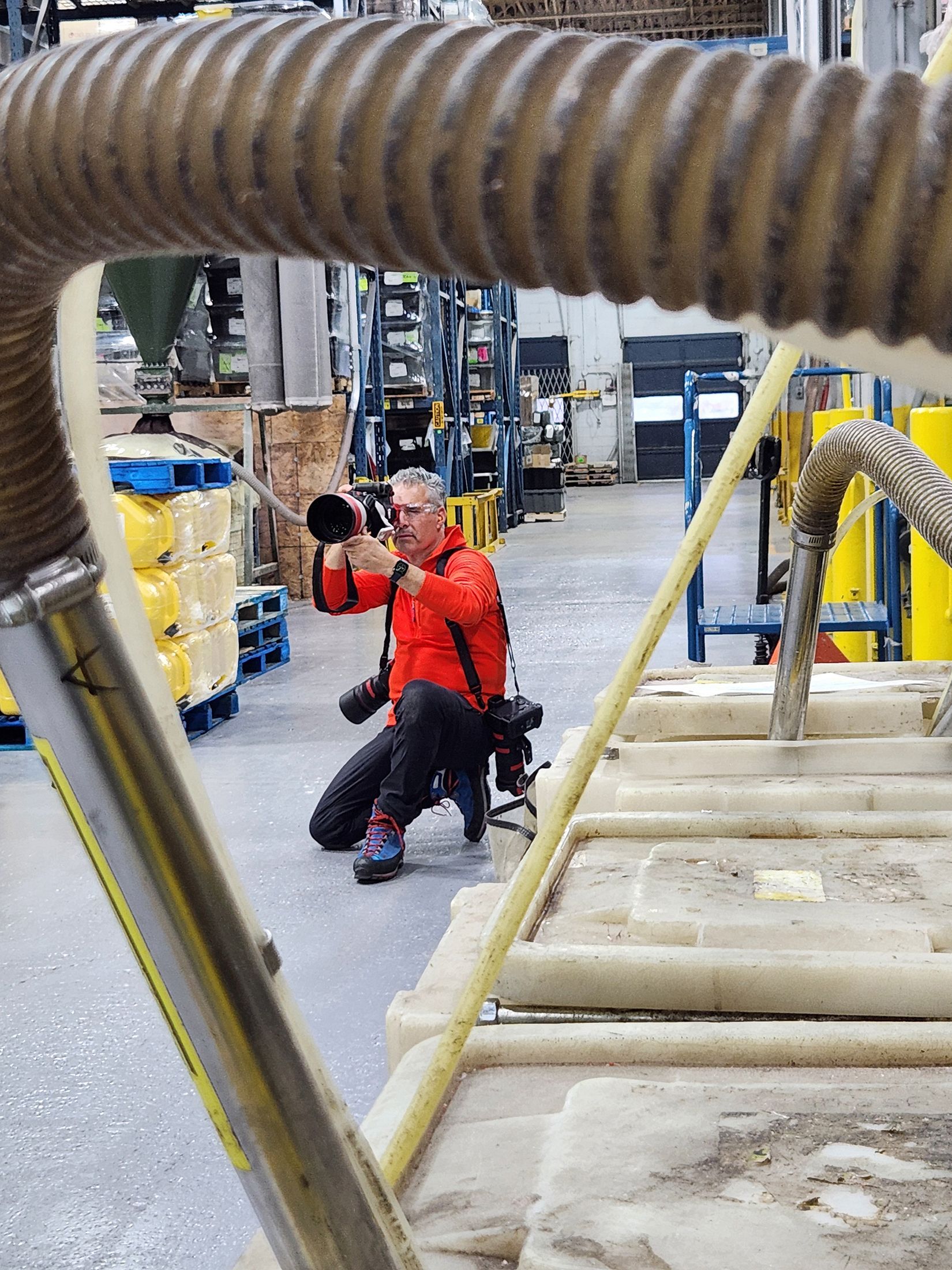 Commercial photographer kneels and holds a large camera lens as he takes photos in a large manufacturing facility.