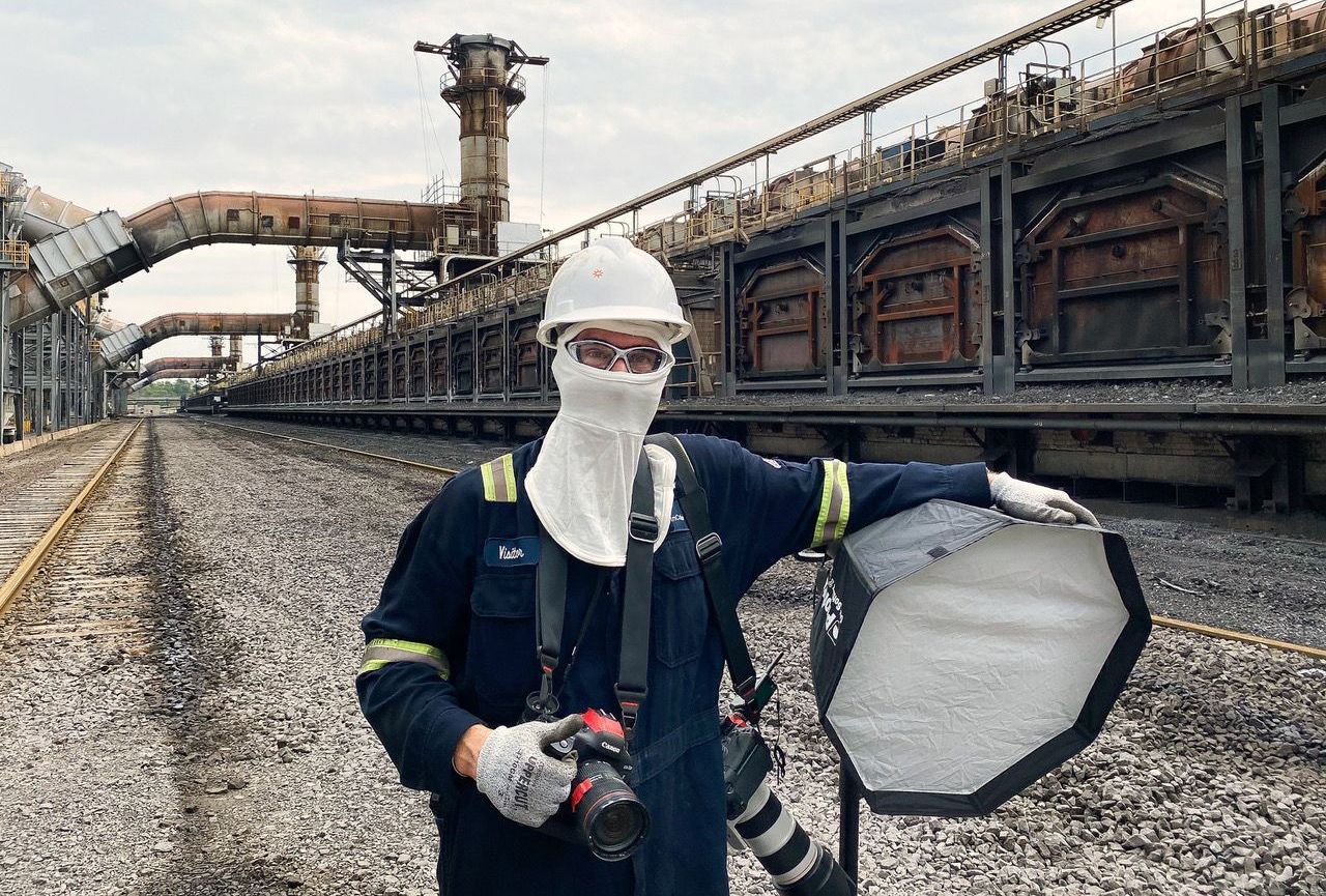 An industrial photographer dressed in protective gear holds his camera and light kit while standing outside of an industrial manufacturing facility.