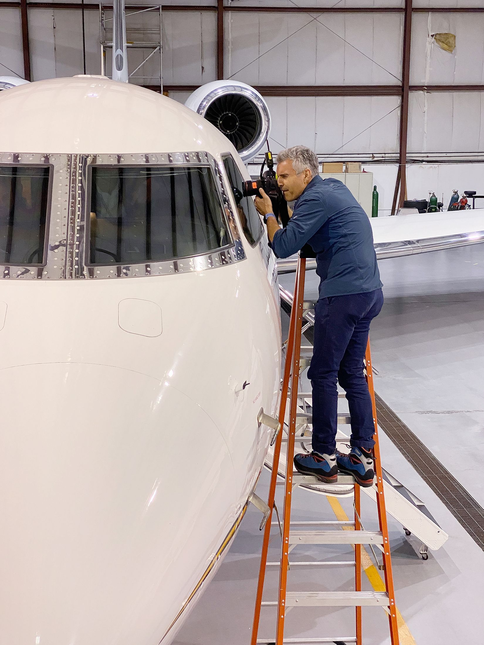 Professional commercial photographer Patrick Schneider takes photos of the cockpit of an airplane that is located in a manufacturing facility. The photographer is outside of the airplane and is looking into the cockpit from the outside.