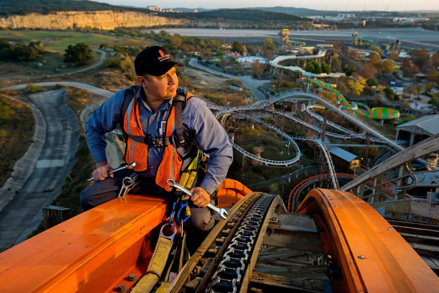 A mechanic is sitting at the top of a roller coaster. The amusement park is shown in the background. This is a portrait of the man at work. He is not looking at the camera. 