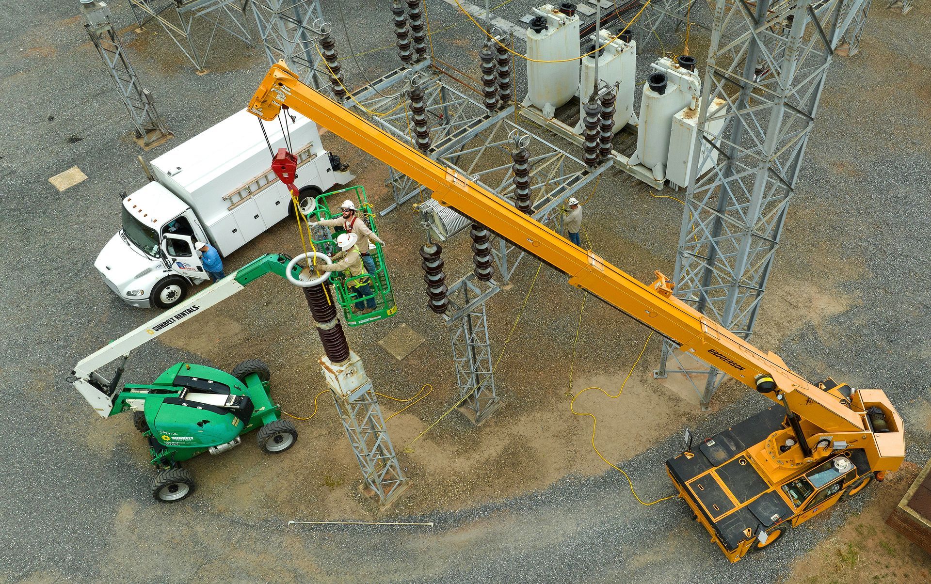 Utility workers standing on a lift about 20 feet above the ground install high-energy power lines. The photo was taken using a drone and the image looks down on the utility workers. 