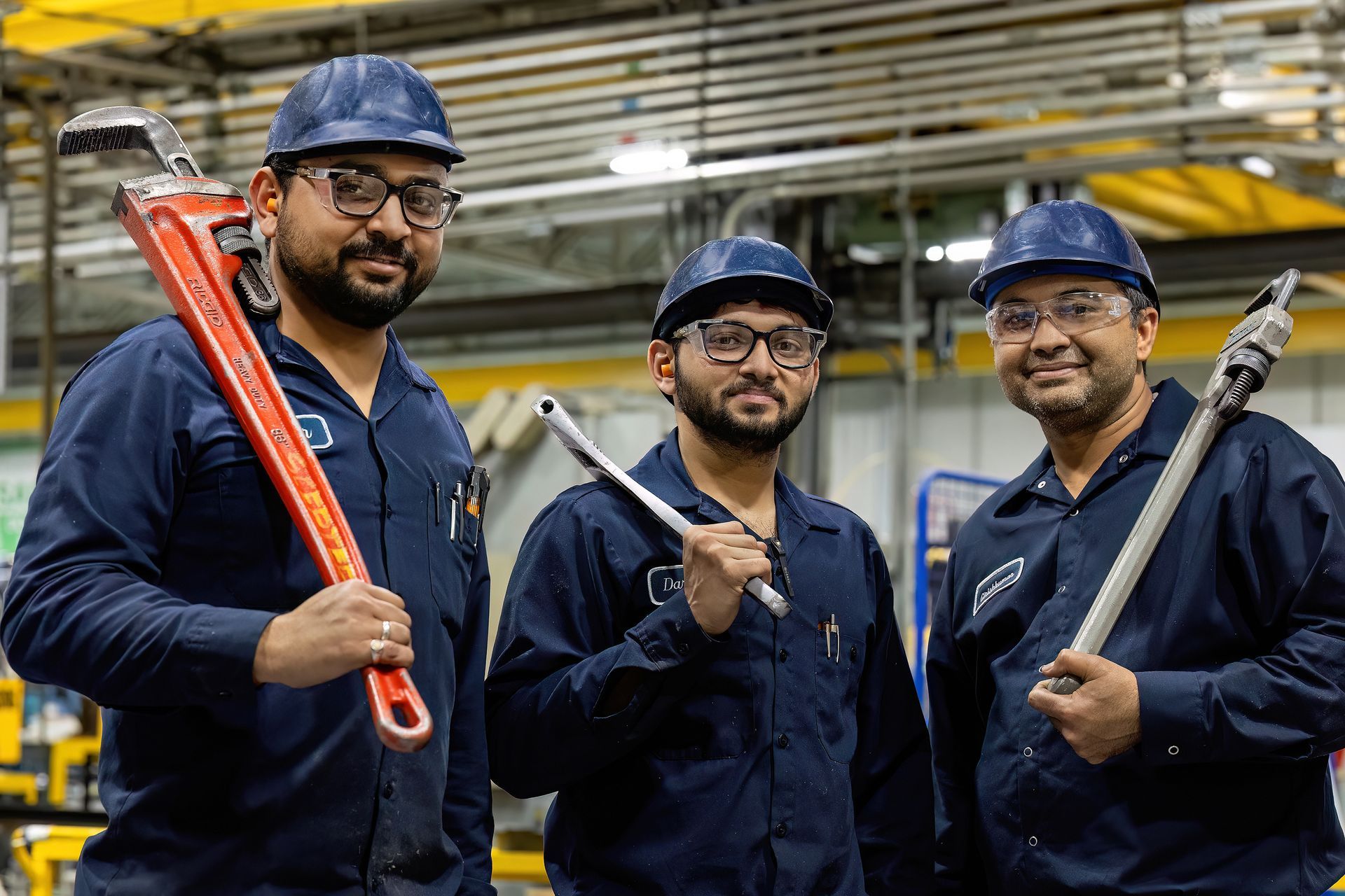 Three workers holding large wrenches smile at the camera. The workers use the wrenches to maintain the manufacturing facility operations. 