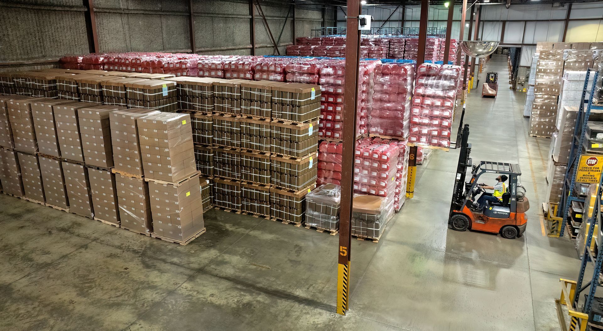 A forklift driver lifts products onto a high shelf in a manufacturing facility. The product is shrink wrapped on a pallet. Photo was made using a drone inside of the facility. 